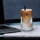 Sheep glass with straw filled with iced coffee and silver straw, placed on a table beside a laptop.