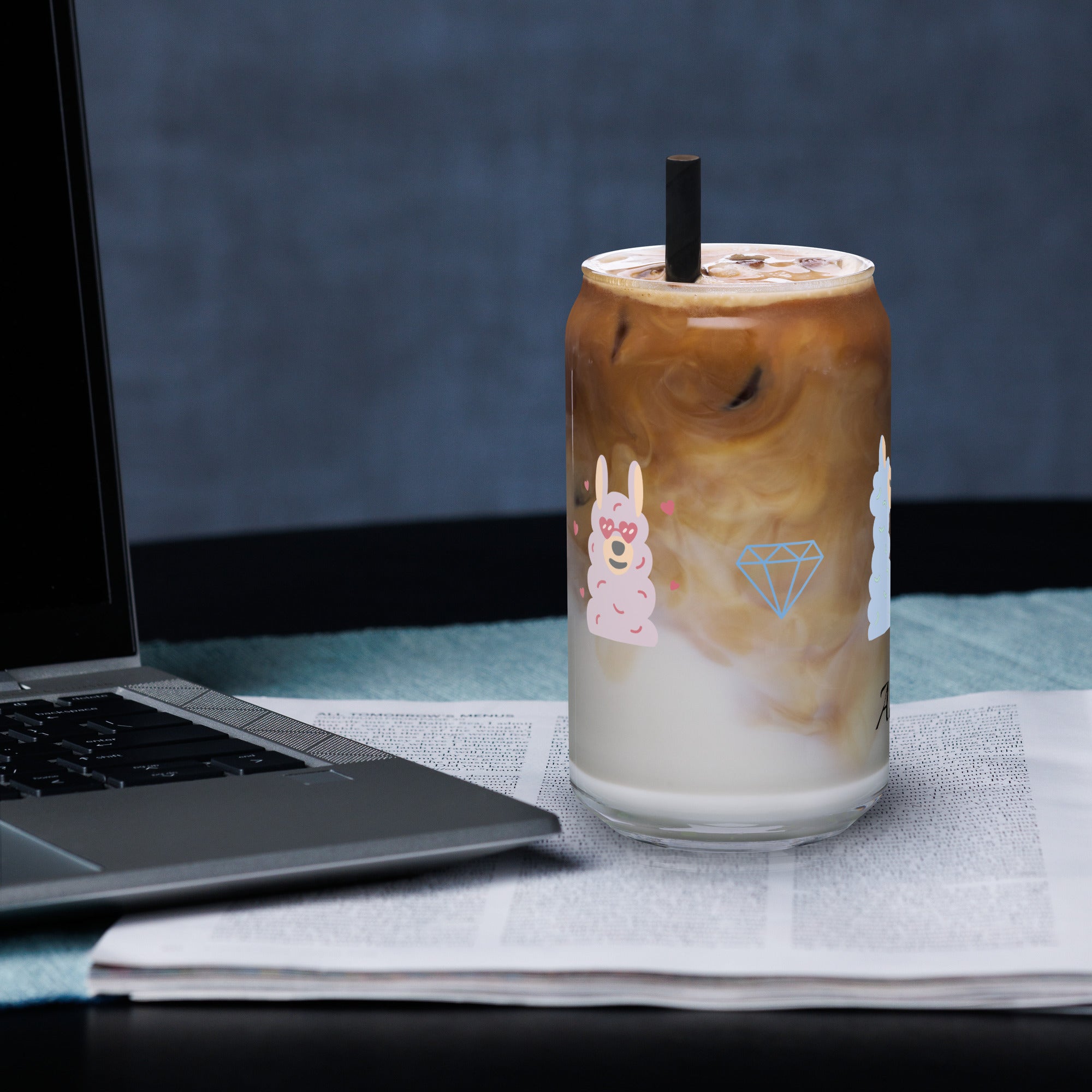 Sheep glass with straw filled with iced coffee and silver straw, placed on a table beside a laptop.