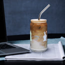 Sheep glass with straw filled with iced coffee and silver straw, placed on a table beside a laptop.